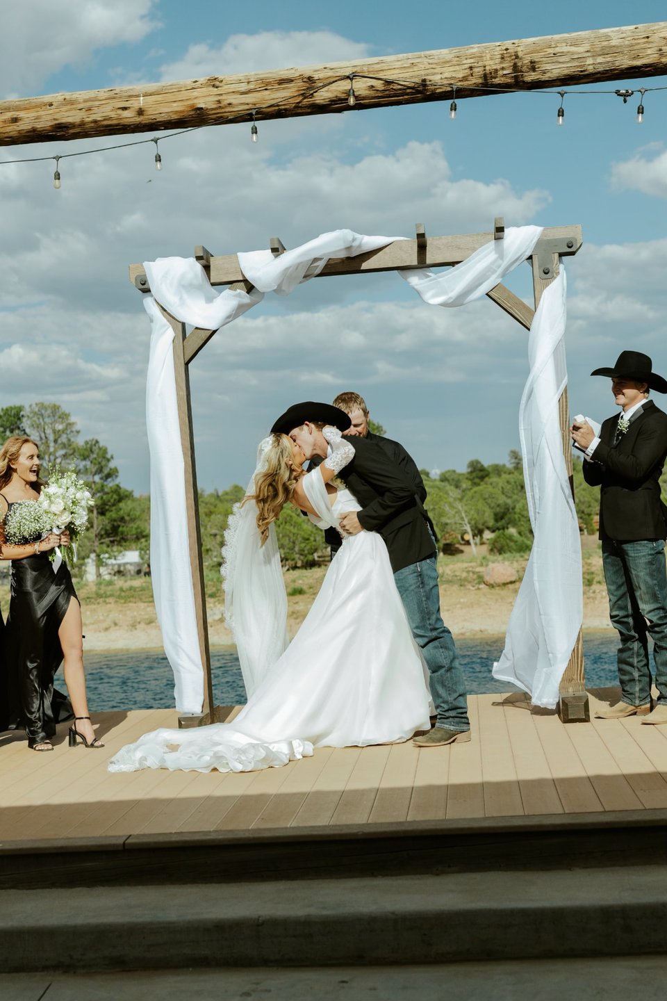 Bride and groom sharing a kiss beneath the wooden wedding arch at a lakeside Arizona ceremony