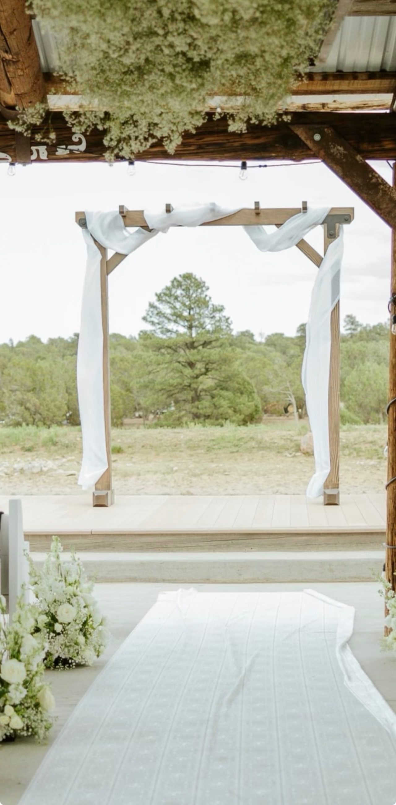 Handcrafted wooden wedding arch draped with white fabric overlooking a floral aisle and white pews in the Arizona pines