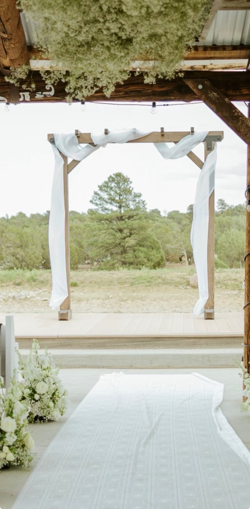 Handcrafted wooden wedding arch draped with white fabric overlooking a floral aisle and white pews in the Arizona pines