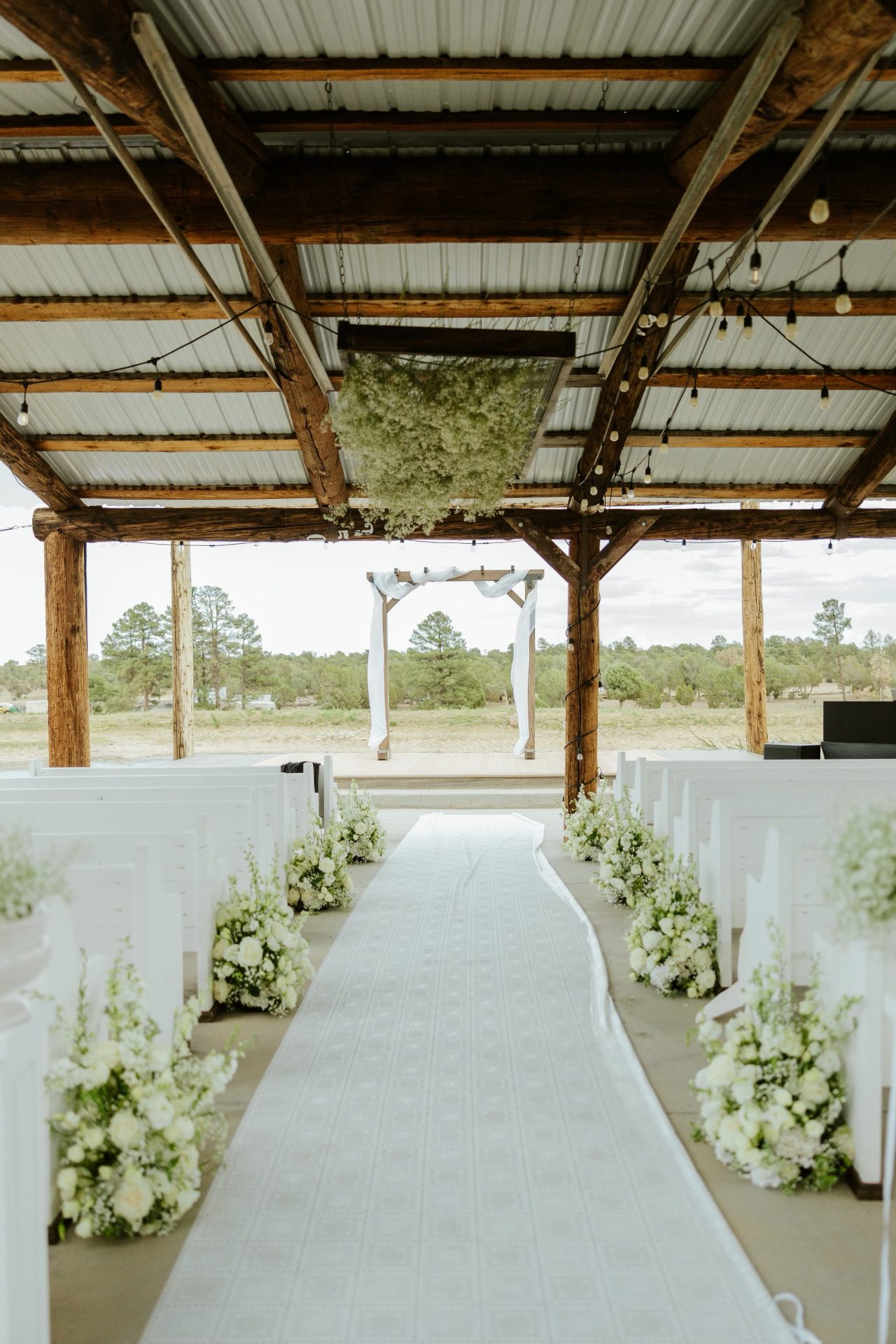 Wooden wedding arch with white draping at a covered White Mountains ceremony aisle lined with white pews and floral arrangements
