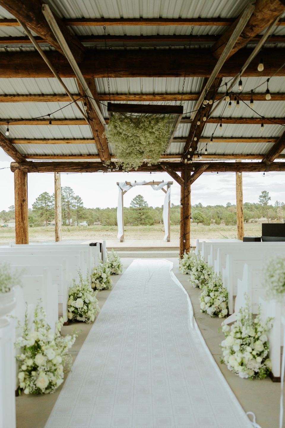 Wooden wedding arch with white draping at a covered White Mountains ceremony aisle lined with white pews and floral arrangements